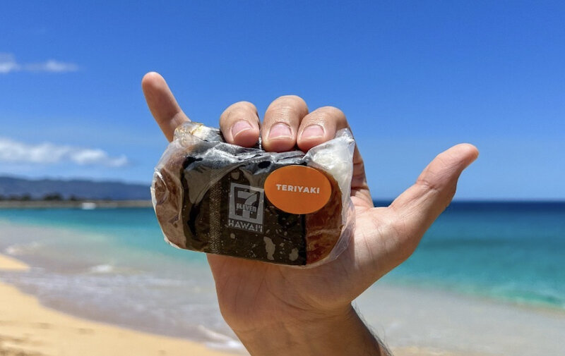 Sunny day at the beach. Person is holding a 7-Eleven teriyaki Spam musubi while making their hand into a shaka.
